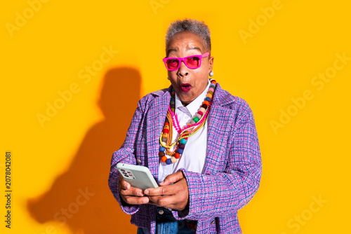 Senior woman reacting with surprise using smartphone - Old hispanic grandmother wearing fashionable modern clothing on colorful background  concepts about elderly  aging  diversity 