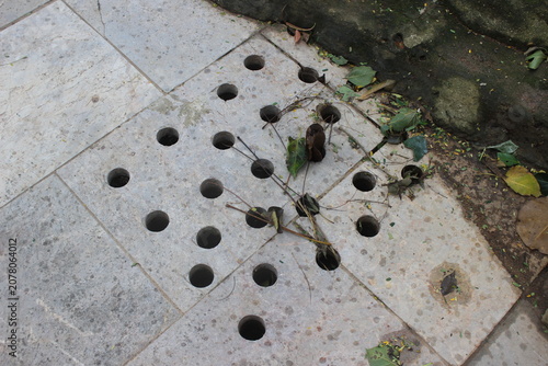 Stone pavement drainage cover with circular holes scattered on textured grey surface.