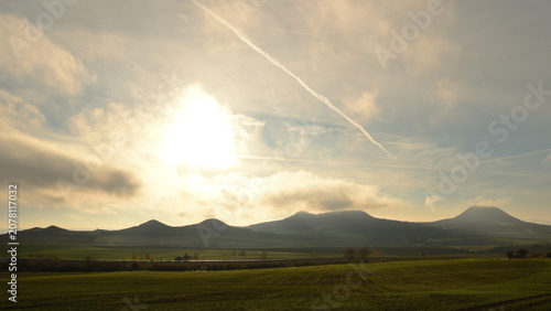 Tableau sur toile Sunlit Volcanic Ridges and Green Fields under a Softly Clouded Sky with Contrail