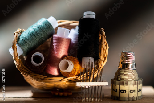 Wicker basket with colorful sewing threads, needles, measuring tape and thimble on a wooden table.