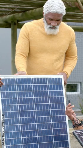 Vertical video: Holding large panel, yellow-sweater man checking fit in yard as tech raising plug