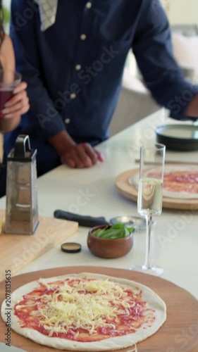 Vertical video: Adding cheese, friends in blue shirt making pizzas in kitchen passing basil to cook