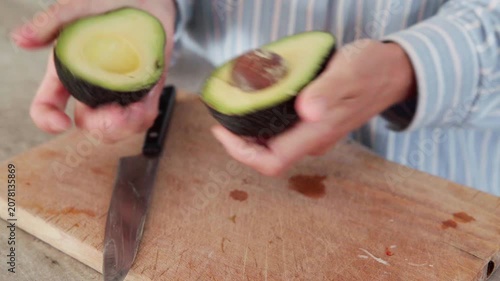 Close up of woman hands opening it in half  and cutting fresh avocado with knife on wooden chopping board in kitchen, preparing healthy organic food recipe for salad at home