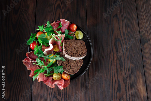 Delicious sliced jamon and bacon with colorful cherry tomatoes and fresh cilantro on a dark plate, rustic wooden background.