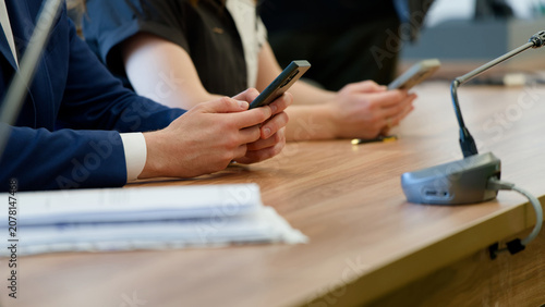 Hands of politicians or lawyers holding mobile devices at a negotiation table. Officials checking messages during a press conference or panel discussion. Corporate setting. Photo