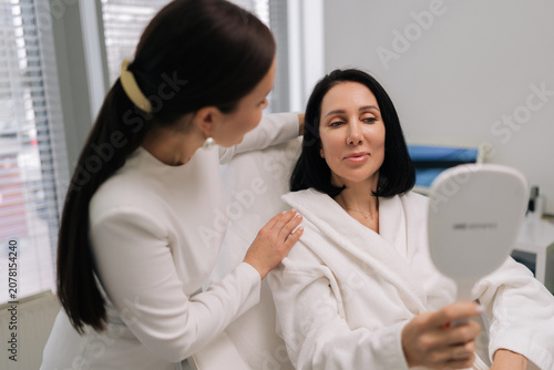 Konstfotografi Friendly beautician doctor providing personalized consultation to female client in specialized treatment chair, reviewing her skin appearance in mirror inside contemporary beauty salon