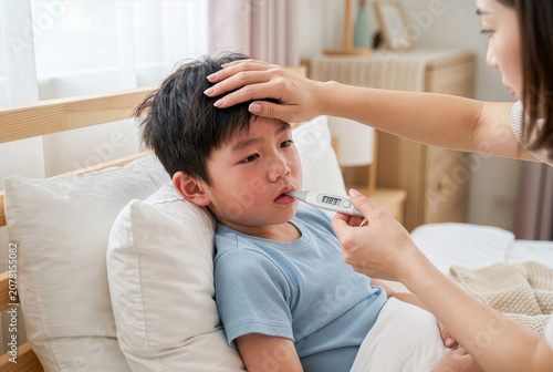 Mother checking her sick little Asian boy's temperature with digital thermometer in bed