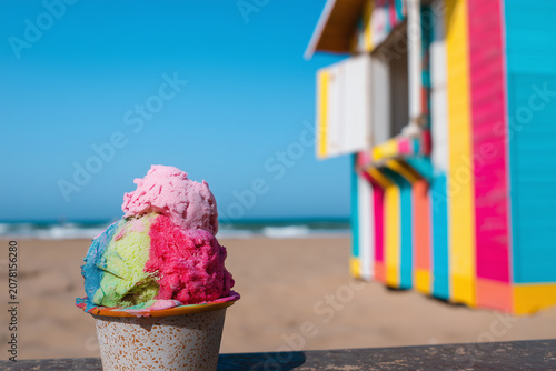 A tub of ice cream scoops in various flavours next to a beach hut on a sunny day during the holidays.