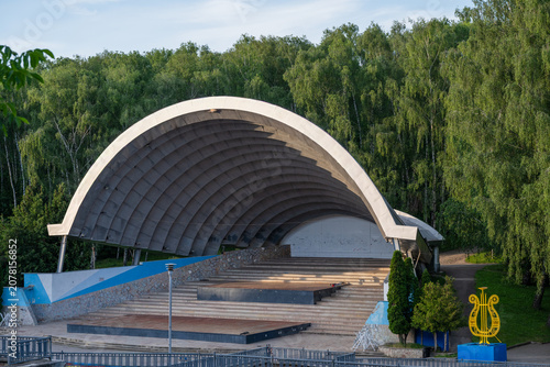 Obraz na płótnie A large, arched concrete bandshell known as Spivoche Pole stands empty, surrounded by lush green foliage in a tranquil public park setting