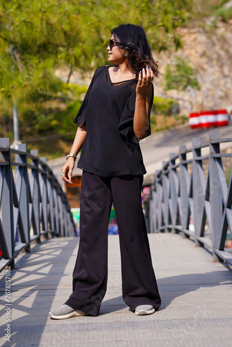 Stylish young woman in black outfit posing on bridge outdoors during sunny day with sunglasses and trendy casual fashion