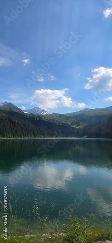A beautiful vertical view of a calm mountain lake perfectly reflecting fluffy white clouds and a bright blue sky, surrounded by dense pine forests and rolling green hills on a sunny day.