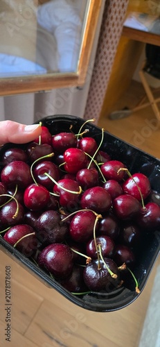 A top-down vertical close-up shot of a hand holding a black plastic container filled with fresh, juicy, and ripe dark red sweet cherries with green stems intact.