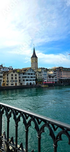 A vertical view captured from a bridge looking across the Limmat River toward traditional European buildings and the historic St. Peter church clock tower under a blue sky in Zurich, Switzerland.