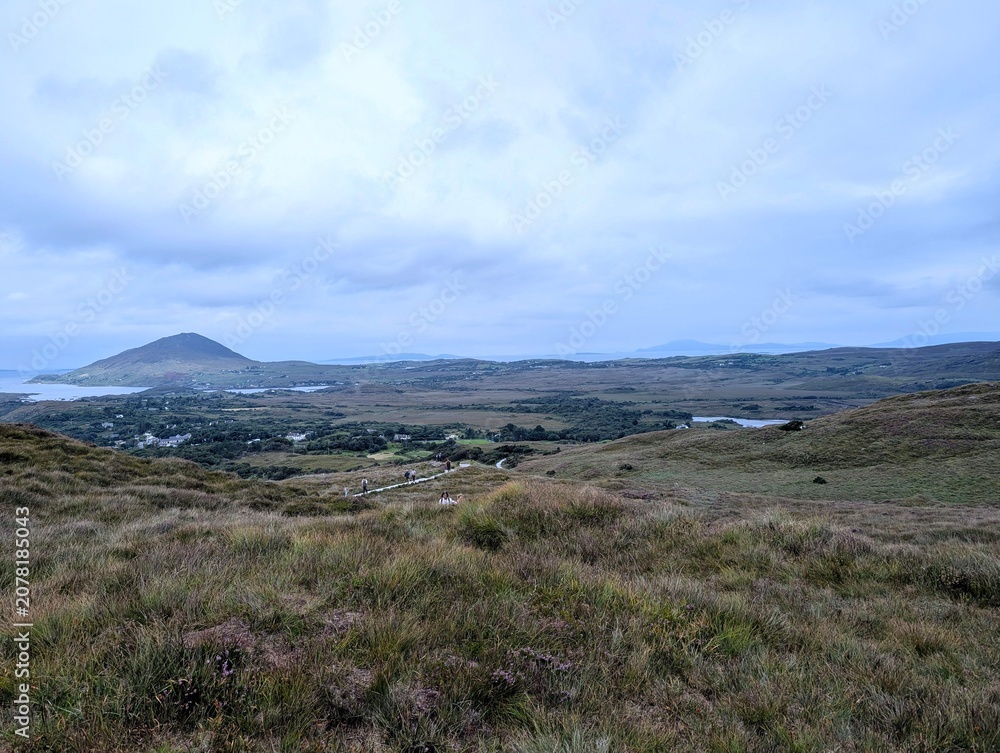 Fototapeta premium Distant hikers walking on a trail through the vast, rugged landscape of Connemara National Park in Ireland.