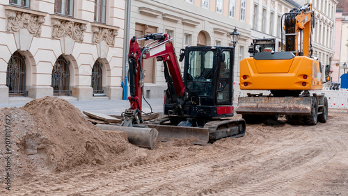 Road construction site in a historic European city street. Repair work with orange plastic pipes, safety barriers, fences, and excavator on the background.