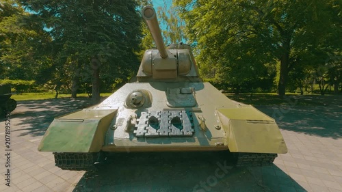 A frontal view of a battered T-34 tank parked on paved ground surrounded by lush green trees. Sunlight casts harsh shadows. The olive-green paintwork shows visible wear