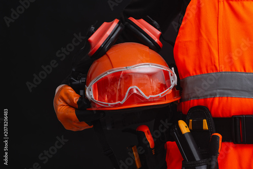 Construction worker holding personal protective equipment including safety helmet, goggles and earmuffs on dark background. Occupational safety, PPE compliance, protection and industrial construction.