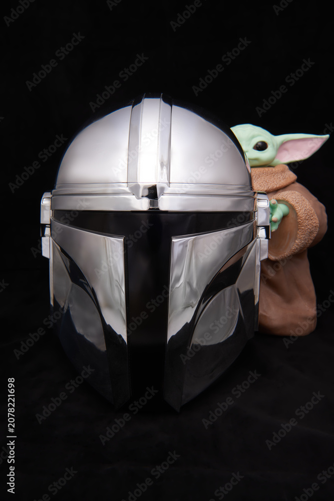 Obraz premium A shiny, metallic Mandalorian helmet on dark velvet, with Grogu holding onto the right side, captured in El Salvador, Cinepolis on June 26, 2026.