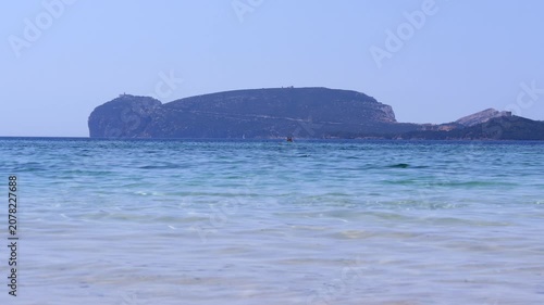 Scenic coastal view of Capo Caccia from Spiaggia di Mugoni, Alghero, Sardinia, featuring lighthouse, calm blue water, gull, and SUP