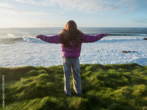Foto Woman stands on grassy cliff near ocean waves