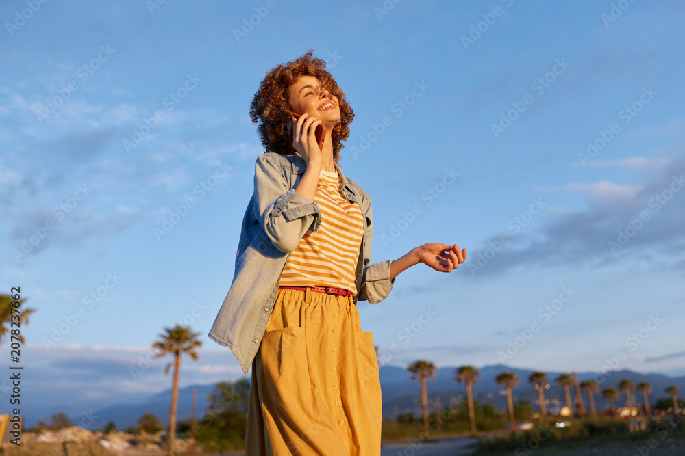 Fototapeta premium Young woman with curly hair stands outdoors in bright sunlight, wearing a striped top and mustard pants, smiling as she enjoys a carefree moment with palm trees in the background.
