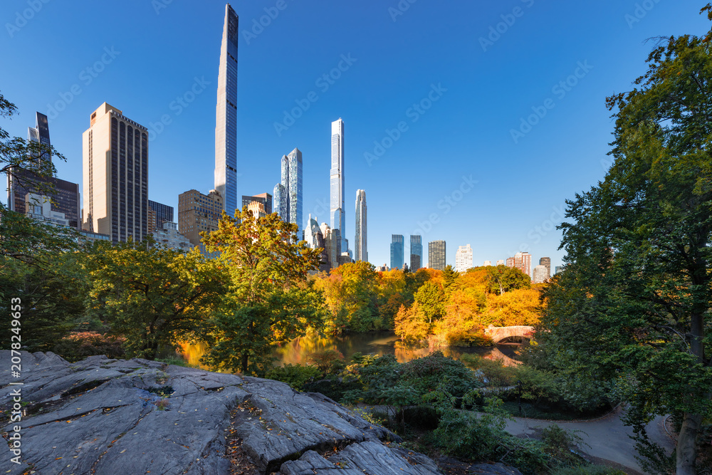 Fototapeta premium Central Park autumn foliage and rock formations with view of Billionaires' Row supertall skyscrapers. Midtown Manhattan, New York City