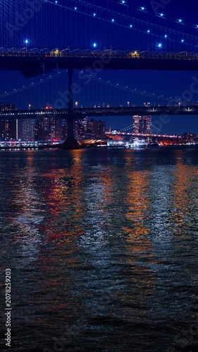 Dark riverscape shimmers with city lights at night. Low angle view at the Manhattan Bridge and the Brooklyn Bridge in New York City. Vertical video.