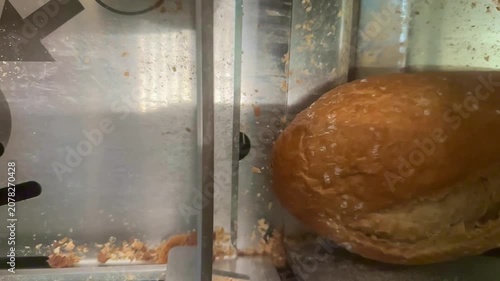 A top-down view shows a mechanical slicer cutting a loaf of artisan bread into uniform slices. The industrial setting features stainless steel surfaces with small crumbs scattered near the blades.