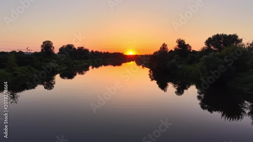 Aerial view of a tranquil river at sunset, showcasing vibrant orange and yellow hues reflecting on the water, surrounded by lush green trees and serene landscape
