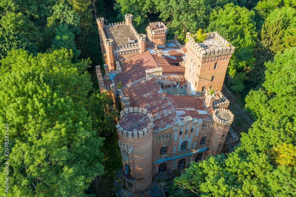 Fototapeta premium Abandoned Neo-Gothic Castle Top-Down Aerial View — Ruined Tudor Manor with Battlements and Round Towers Hidden in Dense Forest | Historic Estate from Above