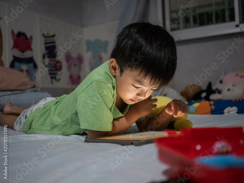 Obraz na płótnie A young boy lies on his bed in a cozy bedroom at night, engrossed in using a tablet
