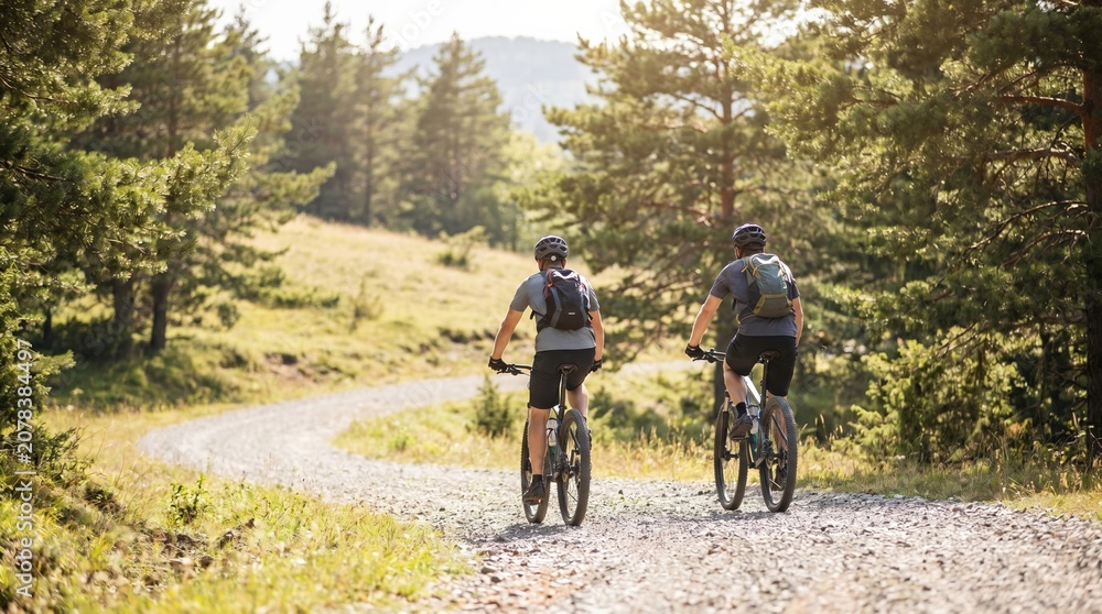 Naklejka premium Two adult men mountain biking on a gravel forest trail during a sunny summer day, active lifestyle and outdoor adventure concept