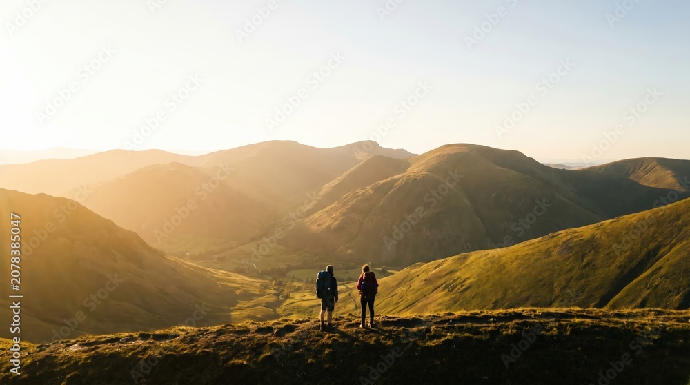 Fototapeta premium Two hikers standing on a mountain ridge overlooking a sunlit valley at sunset, representing adventure, freedom, and outdoor exploration