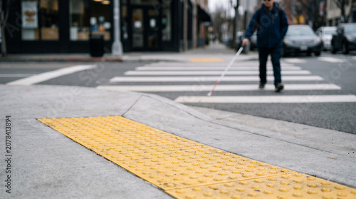 A visually impaired person using a white cane to navigate a city crosswalk with tactile paving on the curb ramp in the foreground