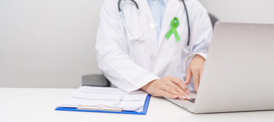 doctor using laptop and report notes on notebook. lime Green Ribbon for concept of May Celiac Awareness month, Lyme disease, September Lymphoma, Mental Health Awareness month and October Liver cancer