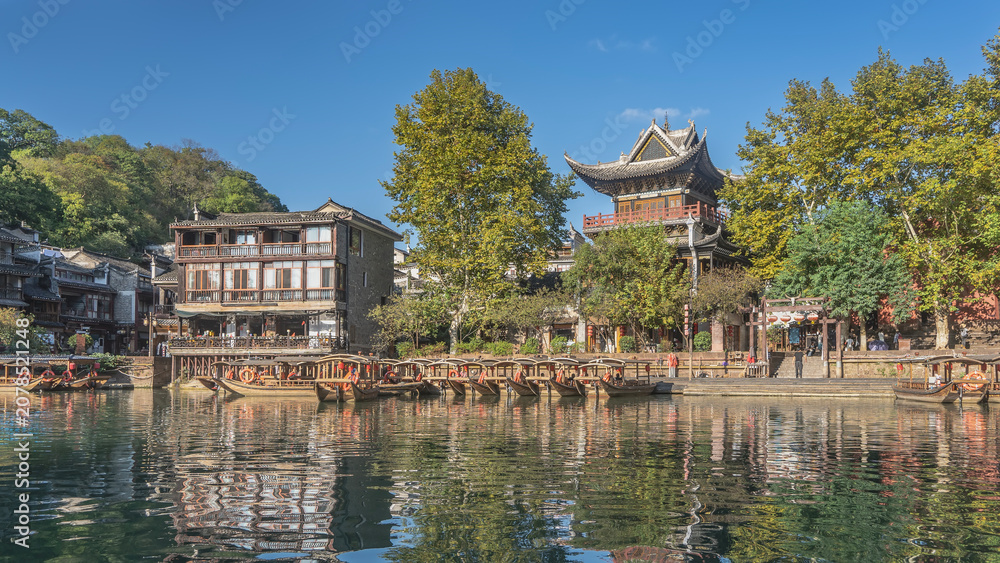 Fototapeta premium Tourist wooden boats with canopies and lifebuoys are moored in a row by the river bank. Reflection. Green trees. Chinese buildings with curved roofs against the blue sky. China. Fenghuang Ancient Town