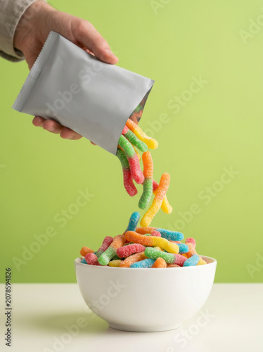 Colorful gummy worms pouring from a silver foil bag into a white bowl, sweet sour candy falling, bright green background studio shot