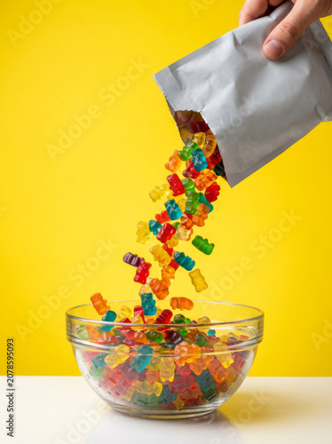 Colorful gummy bears pouring from a silver pouch into a glass bowl, vibrant yellow background, sweet gelatin candy snacks