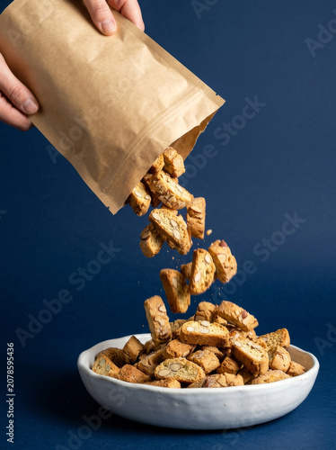 Italian cantucci biscuits pouring from paper bag, crunchy almond biscotti falling into ceramic bowl, sweet bakery dessert on blue background