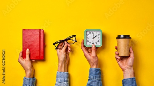 Hands holding book, eyeglasses, alarm clock and coffee cup on yellow background with copy space for productivity concept