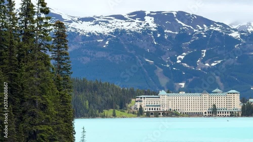 The camera shows a lake with blue water and a historical hotel in the background. Mountains surround the area with some snow on the peaks. Trees line the shore.