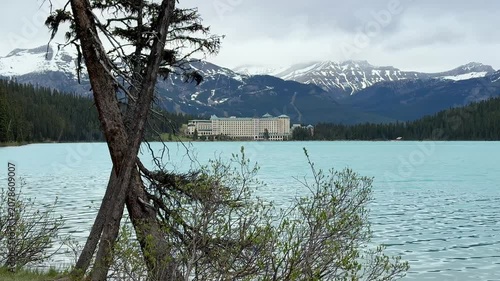 The camera shows a lake with blue water and a historical hotel in the background. Mountains surround the area with some snow on the peaks. Trees line the shore.