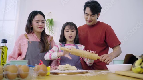 Asian girl flattening raw dough with wooden rolling pin alongside her parents