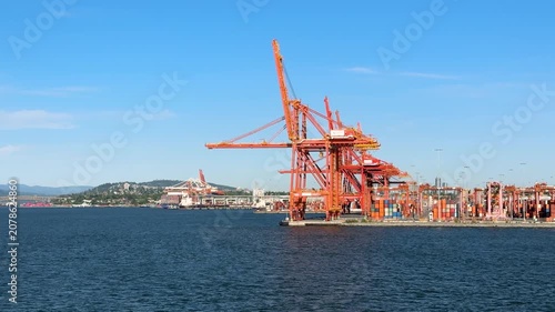 Cranes move containers at a busy port along the water. Ships are docked as workers manage cargo. The sun sets behind the city skyline, adding a vibrant touch to the scene.