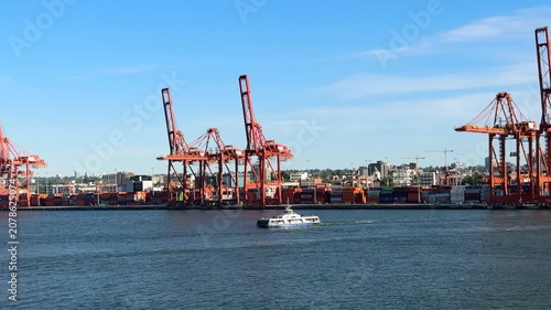 Cranes move containers at a busy port along the water. Ships are docked as workers manage cargo. The sun sets behind the city skyline, adding a vibrant touch to the scene.