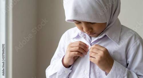 Young muslim schoolgirl in white hijab buttoning her uniform shirt indoors