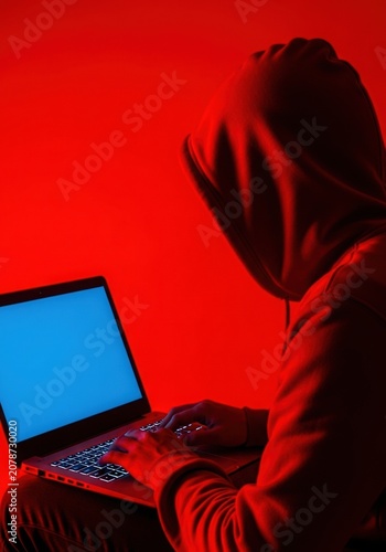Anonymous hacker using laptop under red lighting, person in hooded sweatshirt uses laptop. Red lighting and dark background. Realistic style with strong contrast. Cybersecurity, hacking, cybercrime