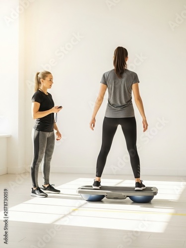 Personal trainer supervising balance exercise session, Female trainer supervises young woman balancing on a stability platform. Bright fitness studio with natural light. Realistic style. Functional