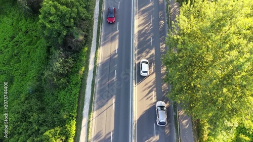 Aerial view of a white car driving along a summer road surrounded by greenery, showcasing the continuous journey over the asphalt and across a bridge above water