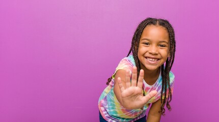 back to school Happy little girl with braided hair wearing a colorful tie dye shirt waving her hand at the camera against a bright solid purple background studio